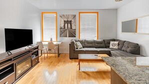 Ein modernes Wohnzimmer mit großem Flachbildfernseher, einer grauen Eckcouch, Holzböden und zwei Fenstern mit Jalousien.