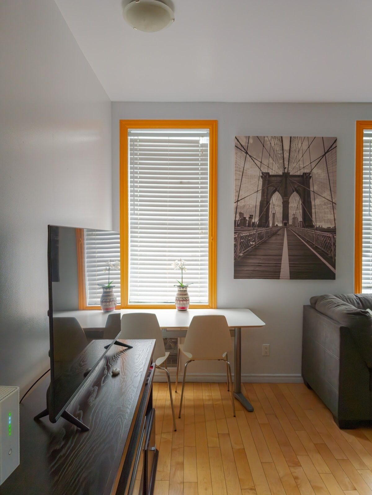 A modern living room with a dining area, wooden flooring, a flat-screen TV, and a large window with blinds.