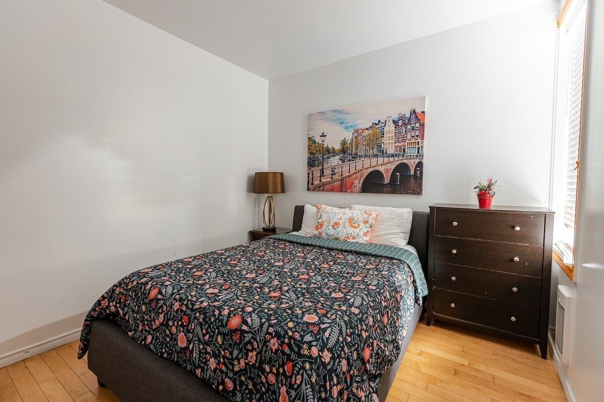Um quarto com cama, cômoda, abajur e pintura de uma ponte.