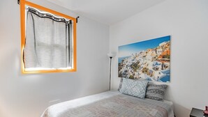 Ein Schlafzimmer mit einem Bett, einem Fenster mit Vorhängen, einer Lampe und einem Bild von einem Küstendorf.