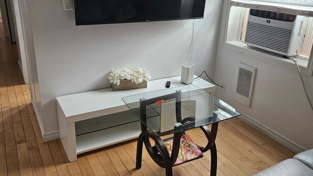 Ein Wohnzimmer mit Flachbildfernseher an der Wand, einem Glastisch, einer weißen Konsole und einem grauen Sofa.