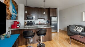 Een moderne keuken met donkerhouten kastjes, een granieten aanrechtblad en roestvrijstalen apparatuur. Er staan twee zwarte barkrukken bij het eiland, een grijze bank en een blauwe tafel met een rode koffiezetapparaat.