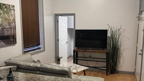 Een ruimtelijk ingedeeld woongedeelte met een keuken, een zithoek met een bank en poef, een televisie op een houten standaard, en een potplant.