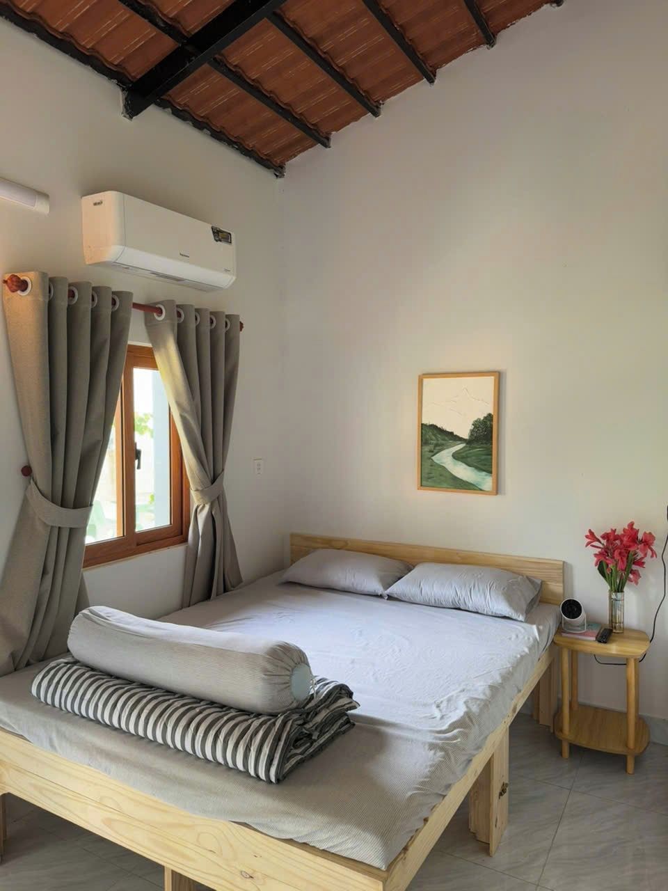 Een slaapkamer met een houten bed, grijze gordijnen, een klein nachtkastje met een bloemenvaas en een schilderij aan de muur.