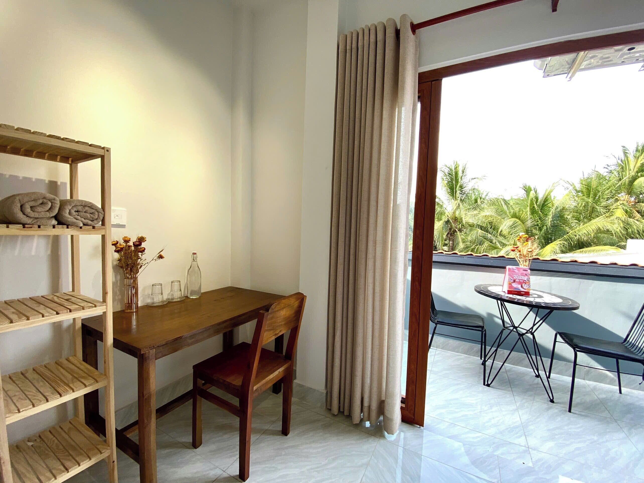 Una habitación con una mesa de madera, una silla, un estante con toallas y vistas a palmeras a través de una ventana con cortinas.