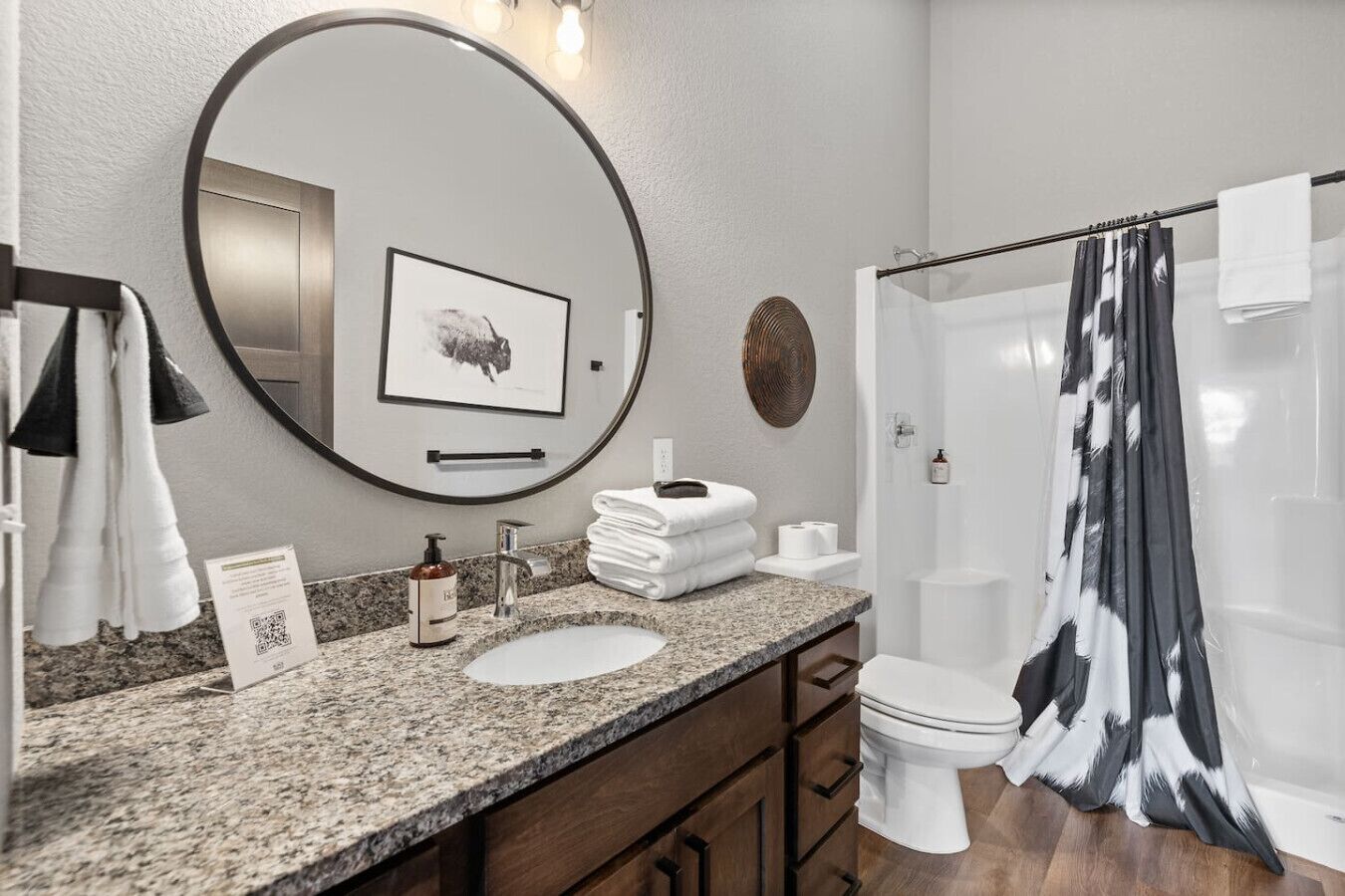 A bathroom with a round mirror, a sink, a toilet, and a shower with a black and white curtain.