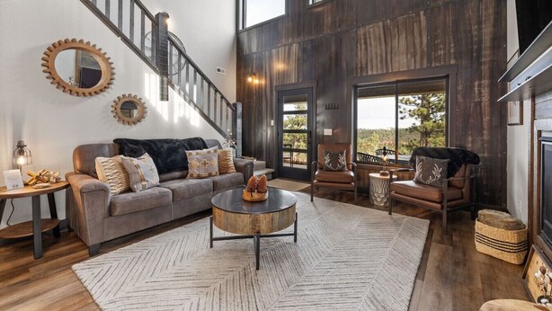 A spacious living room with a large sofa, coffee table, and fireplace. A staircase with a metal railing leads to a balcony with a view of trees. - MK Hideaway Mountain Cabin w/ Hot Tub (Lead)