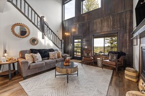 A spacious living room with a large sofa, coffee table, and fireplace. A staircase with a metal railing leads to a balcony with a view of trees. - MK Hideaway Mountain Cabin w/ Hot Tub (Lead)