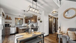 A modern kitchen with stainless steel appliances, a dining area with a wooden table and bench, and a cozy living space with a leather couch and a round mirror on the wall. - MK Hideaway Mountain Cabin w/ Hot Tub (Lead)