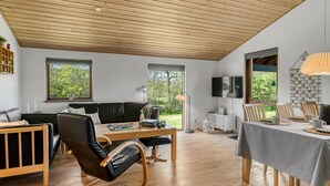 Un salon moderne avec un plafond en bois, un canapé en cuir noir, des chaises en bois, une table à manger recouverte d’une nappe grise et un téléviseur fixé au mur.