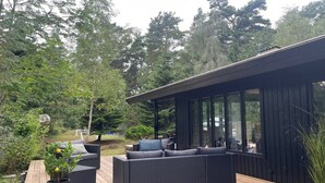 A modern outdoor deck with a sofa and a dining table, surrounded by lush greenery and a wooden deck. - "Sheyla" - 70m from the sea by Interhome (Væggerløse)