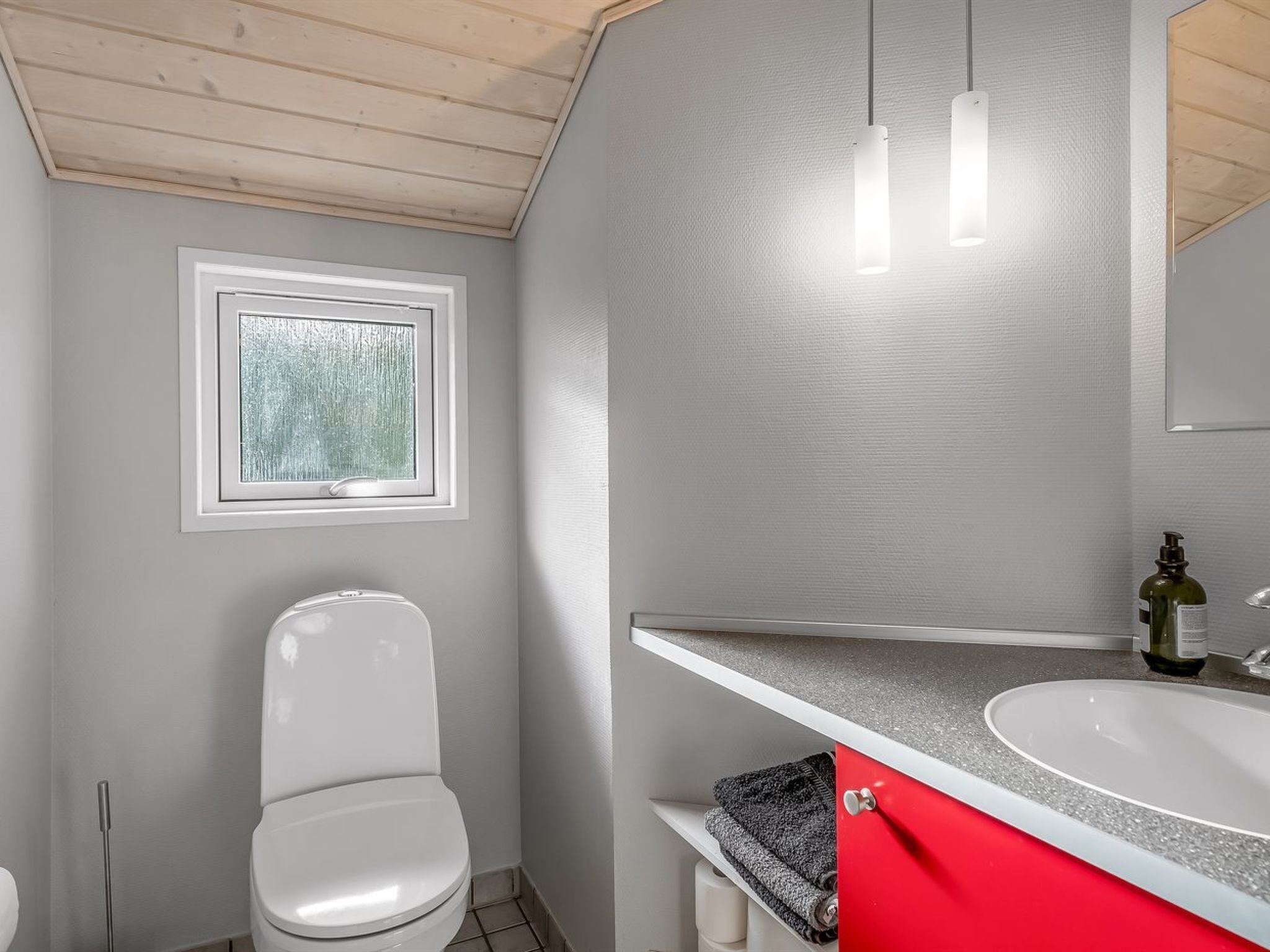 A bathroom with a white toilet, a window, a mirror, and a sink with a red cabinet.