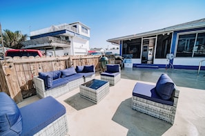 A patio area with wicker furniture and cushions, a wooden fence, and a building with large windows in the background. - Waterfront Pool House on Hutchinson Island (Fort Pierce)