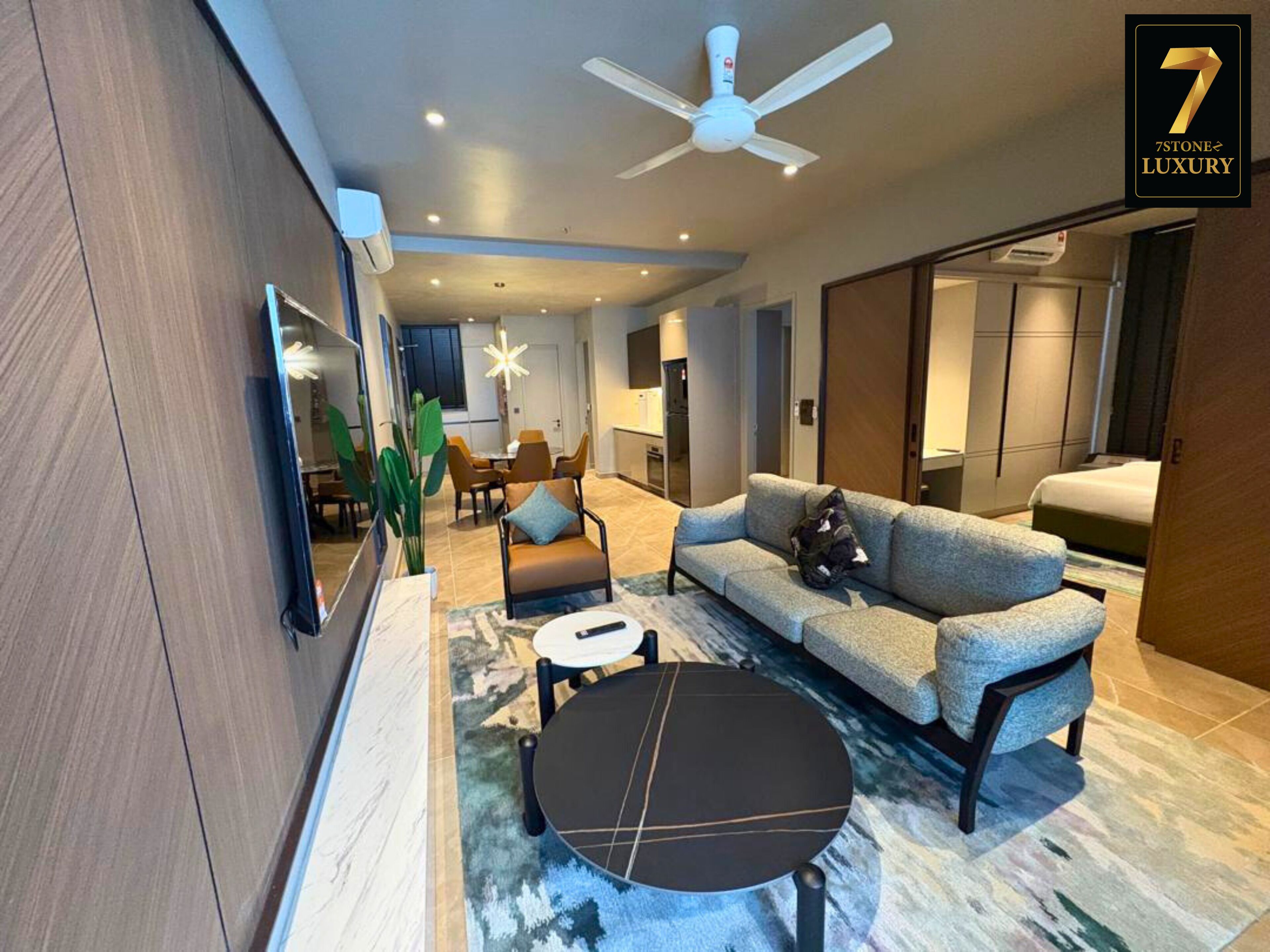 A modern living room with a sofa, coffee table, and a ceiling fan. There is a dining area in the background.