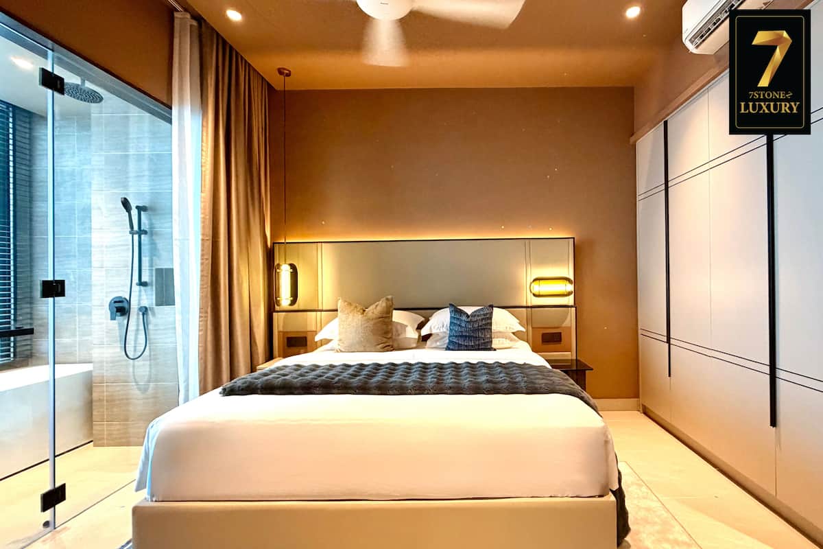 A modern hotel room with a large bed, a glass shower enclosure, and a sign indicating '7 Stone Luxury'.