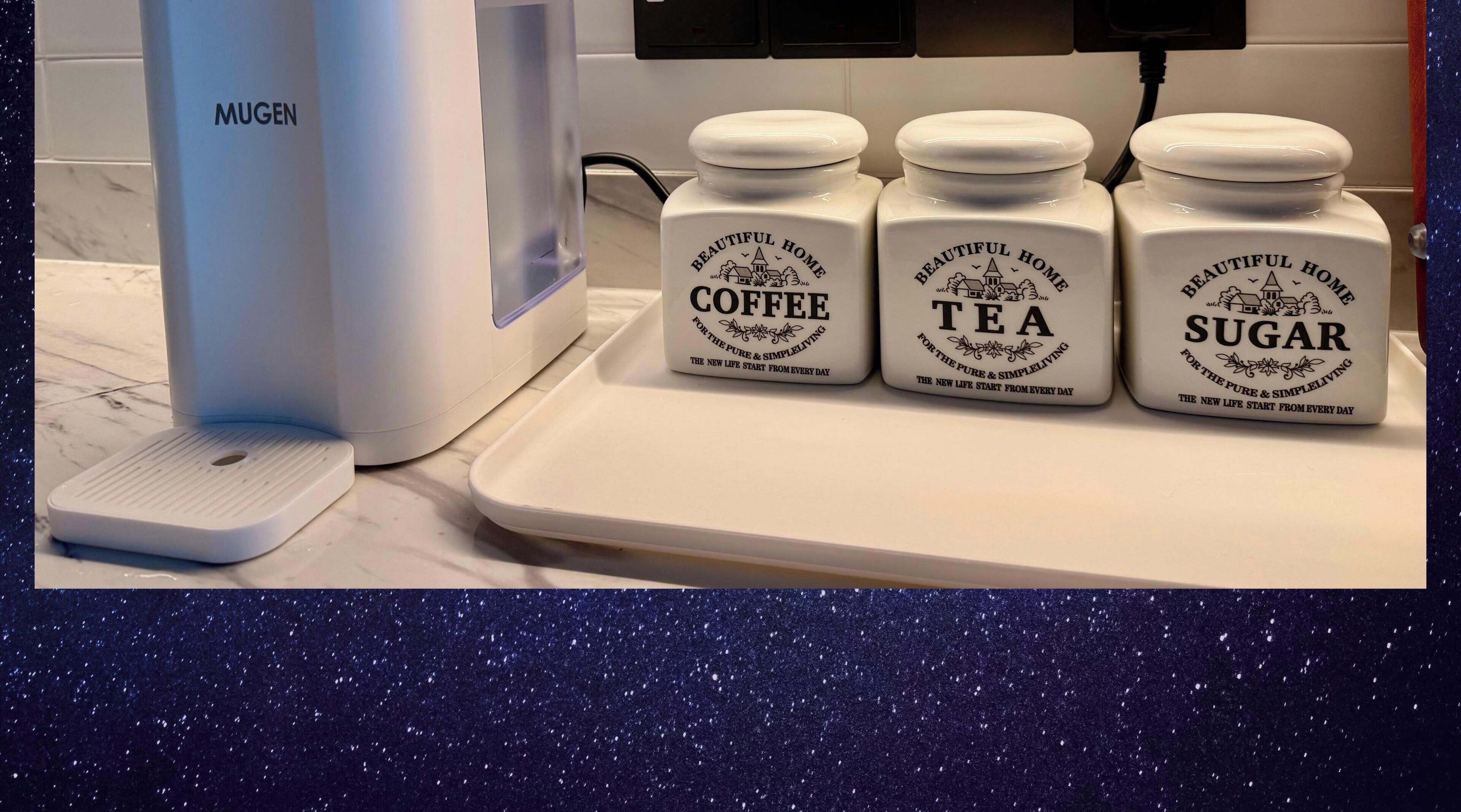 A kitchen counter with a coffee maker, three jars labeled coffee, tea, and sugar, and a scale.