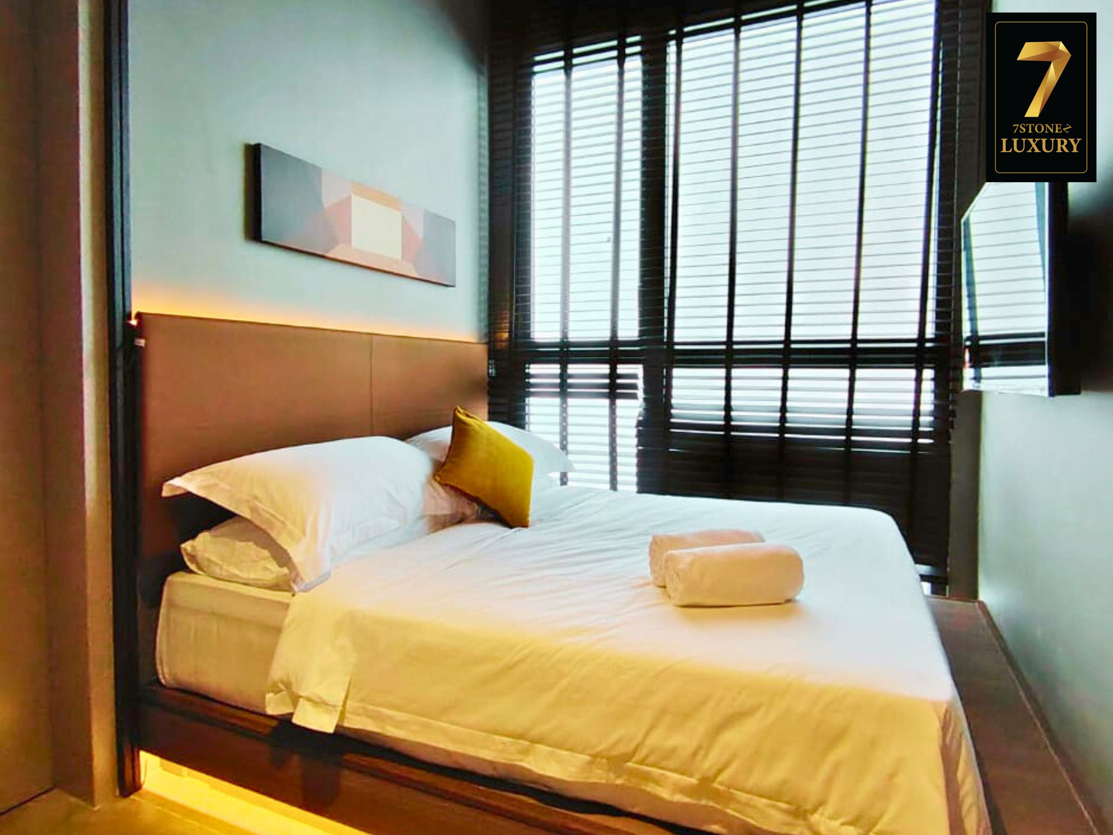 A neatly made bed with white and yellow pillows, a bedside table with a white towel, and a window with blinds.