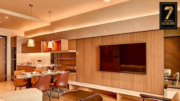 A modern living room with a sofa, coffee table, and TV. There is a dining area with chairs and a table. The room has wooden paneling and a large window.