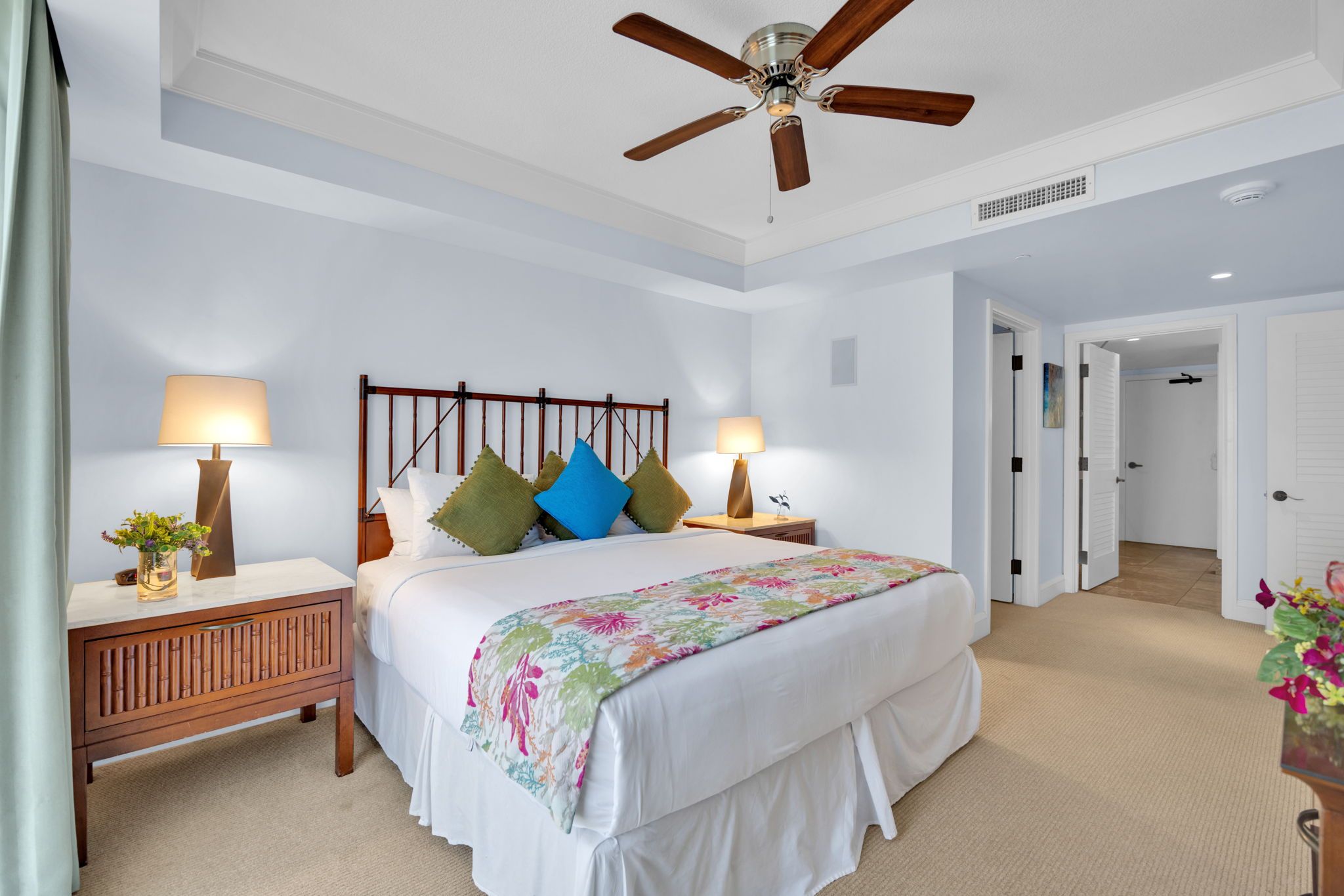 A bedroom with a bed, two bedside tables, a ceiling fan, and a door leading to another room.