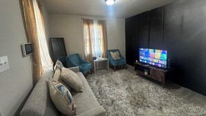 Un salon avec un canapé, un fauteuil turquoise, un téléviseur posé sur un meuble en bois et un luminaire au plafond.