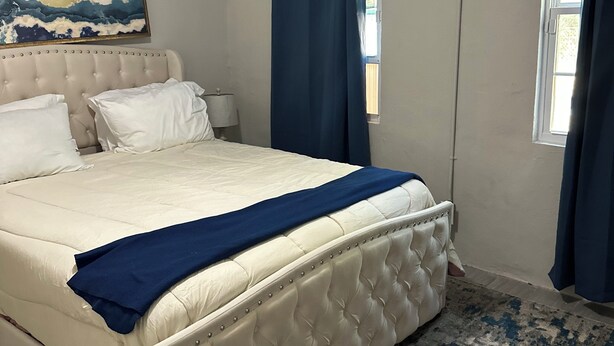 Une chambre à coucher avec un grand lit, une table de chevet, une lampe, un tableau au mur et une fenêtre avec des rideaux.
