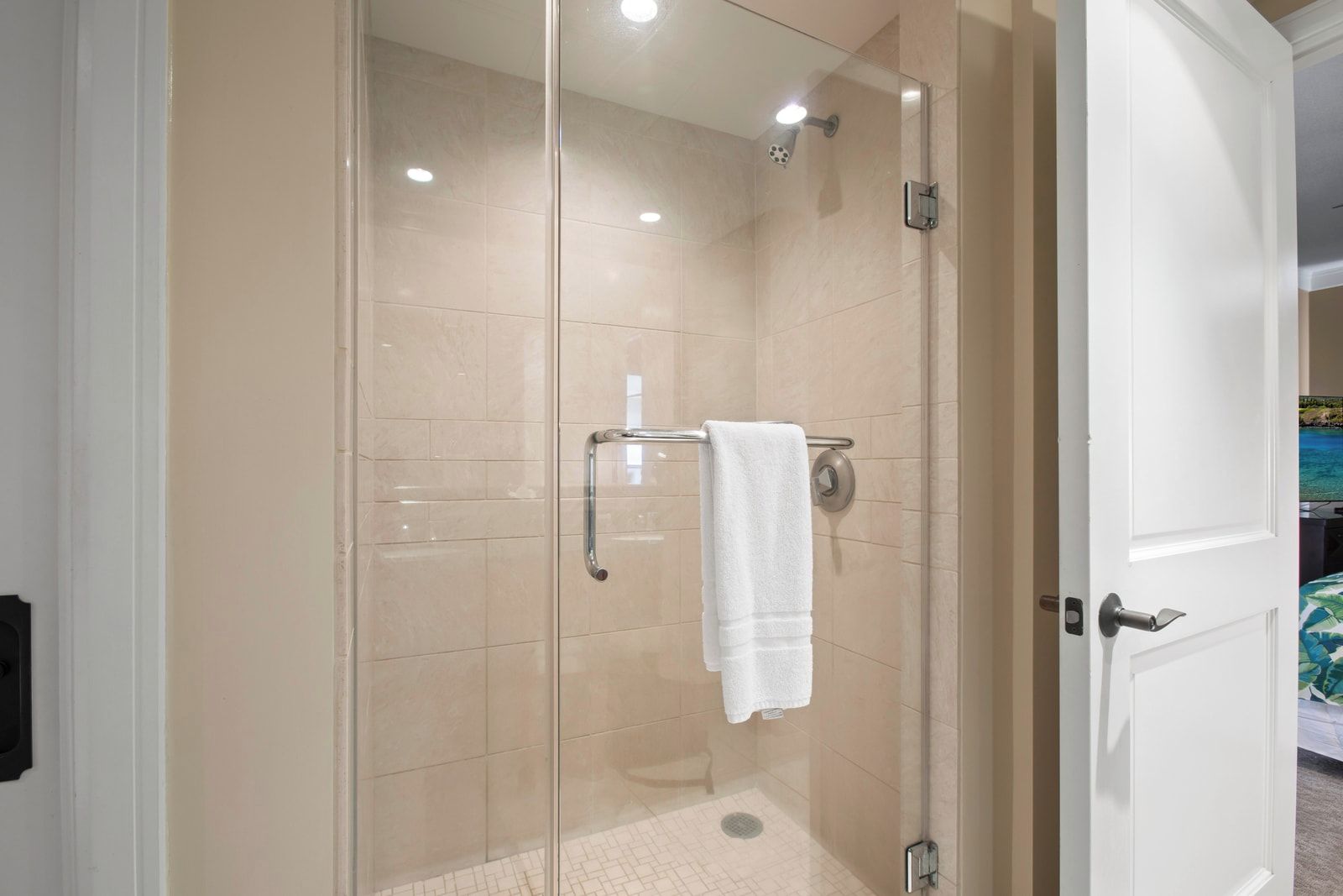 Eine begehbare Dusche mit Glaswänden, ein weißes Handtuch auf einem Metallgestell, ein sichtbarer Türgriff und ein gefliester Boden.