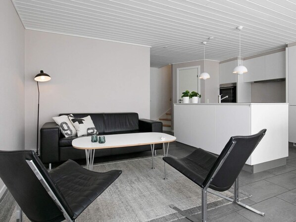 A modern living room with a black leather sofa, a white coffee table, and black chairs. There is a kitchen area with white cabinets and a countertop. - 8 Person Holiday Home on a Holiday Park in Lokken (Lokken)