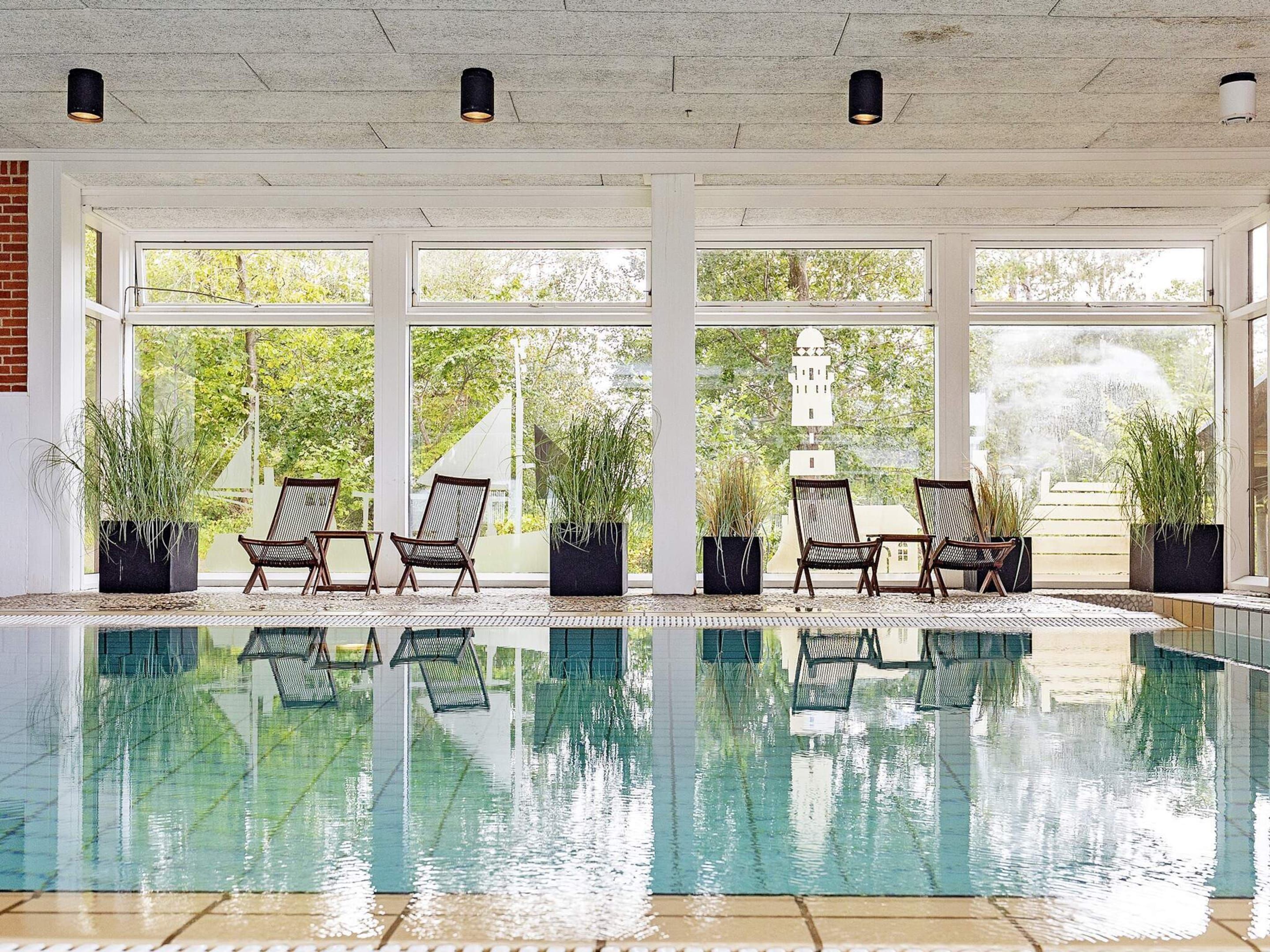Una piscina interior con paredes de vidrio transparente, rodeada de sillones de descanso y plantas en macetas.