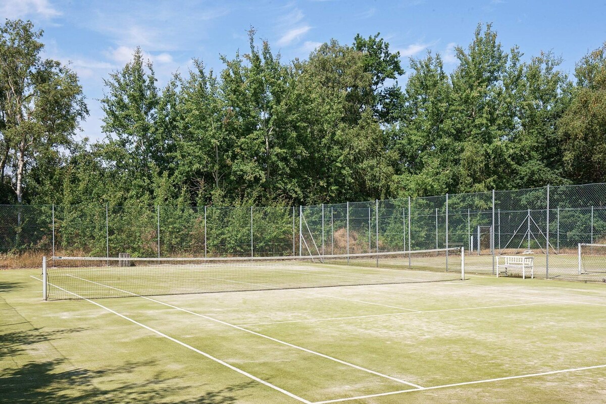An outdoor tennis court with a net, surrounded by a chain-link fence and trees.