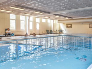 Una piscina interior con techo azulejado, ventanas amplias y algunas sillas de descanso.