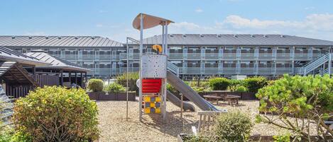 A playground with a slide and climbing structures, surrounded by wooden fences and greenery, with a modern building in the background.