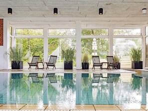An indoor swimming pool with clear glass walls, surrounded by potted plants and wicker chairs.