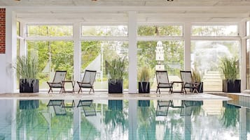 Una piscina interior con paredes de vidrio transparente, rodeada de sillones de descanso y plantas en macetas.