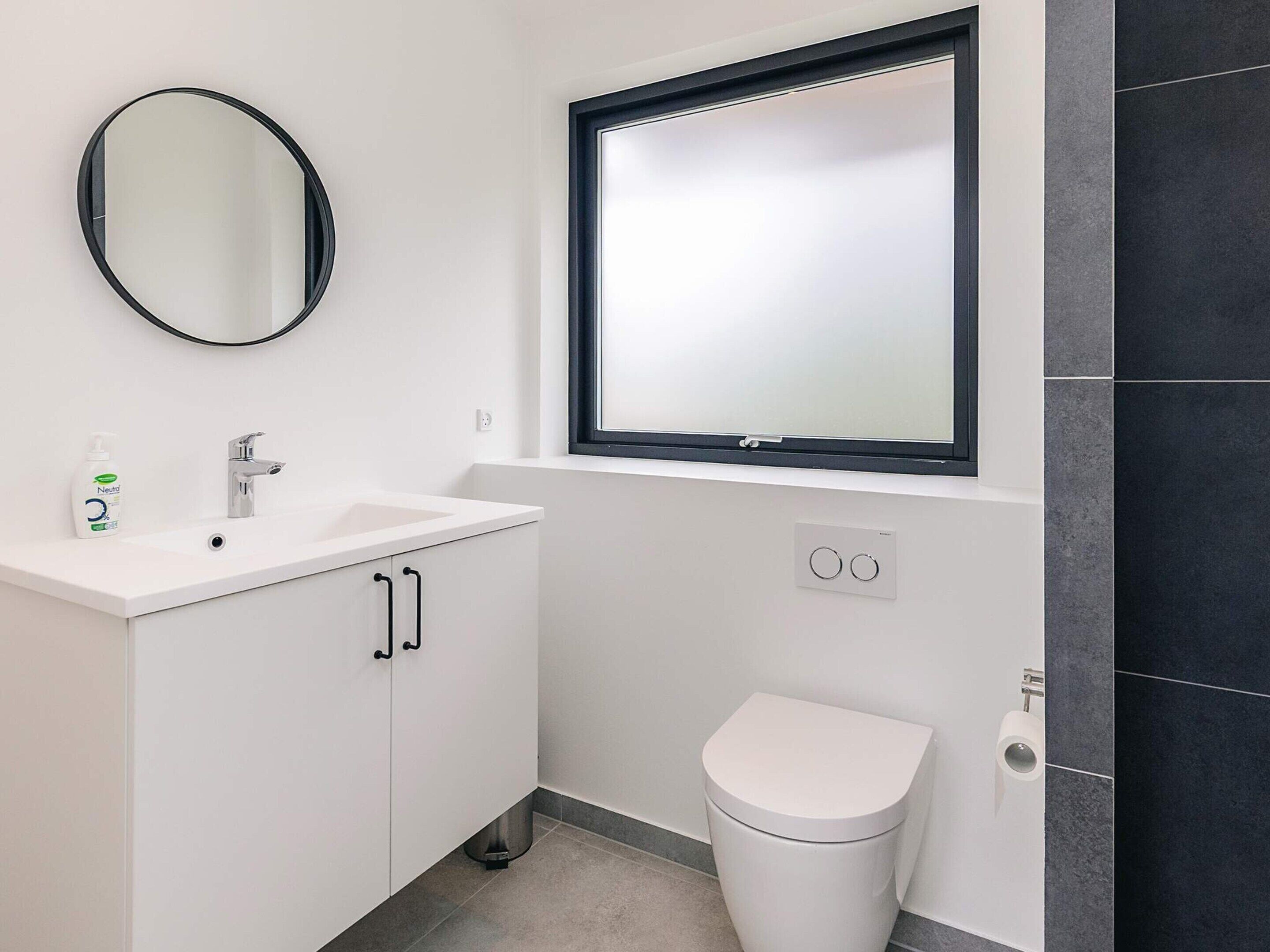 A modern bathroom with a white sink, a round mirror, a toilet, and a window above the toilet.