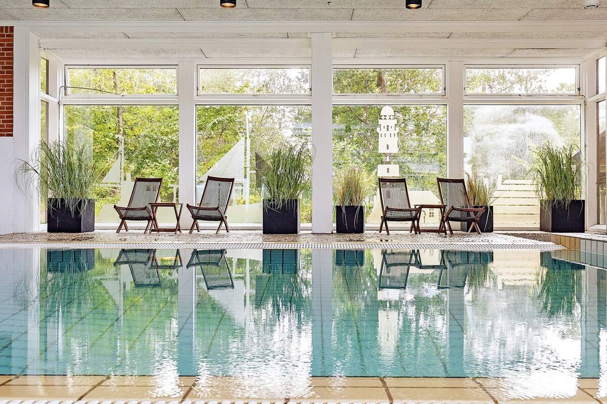 An indoor swimming pool with clear glass walls, two lounge chairs, and potted plants.