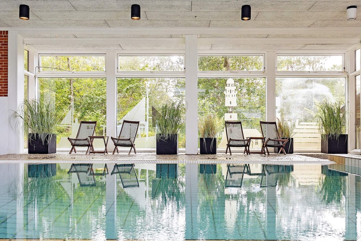An indoor swimming pool with clear glass walls, surrounded by lounge chairs and potted plants.