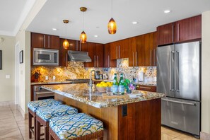 A modern kitchen with wooden cabinets, stainless steel appliances, a granite countertop, and a central island with bar stools. - Honua Kai | Ocean View 5 BR, Sleeps 16 | Car Incl. w/ 6+ Nights | HKH ML-1910 by KBM (Lahaina)