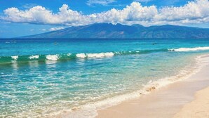 Ein Strand mit klarem türkisblauem Wasser, einem Sandufer und einer Bergkette im Hintergrund.