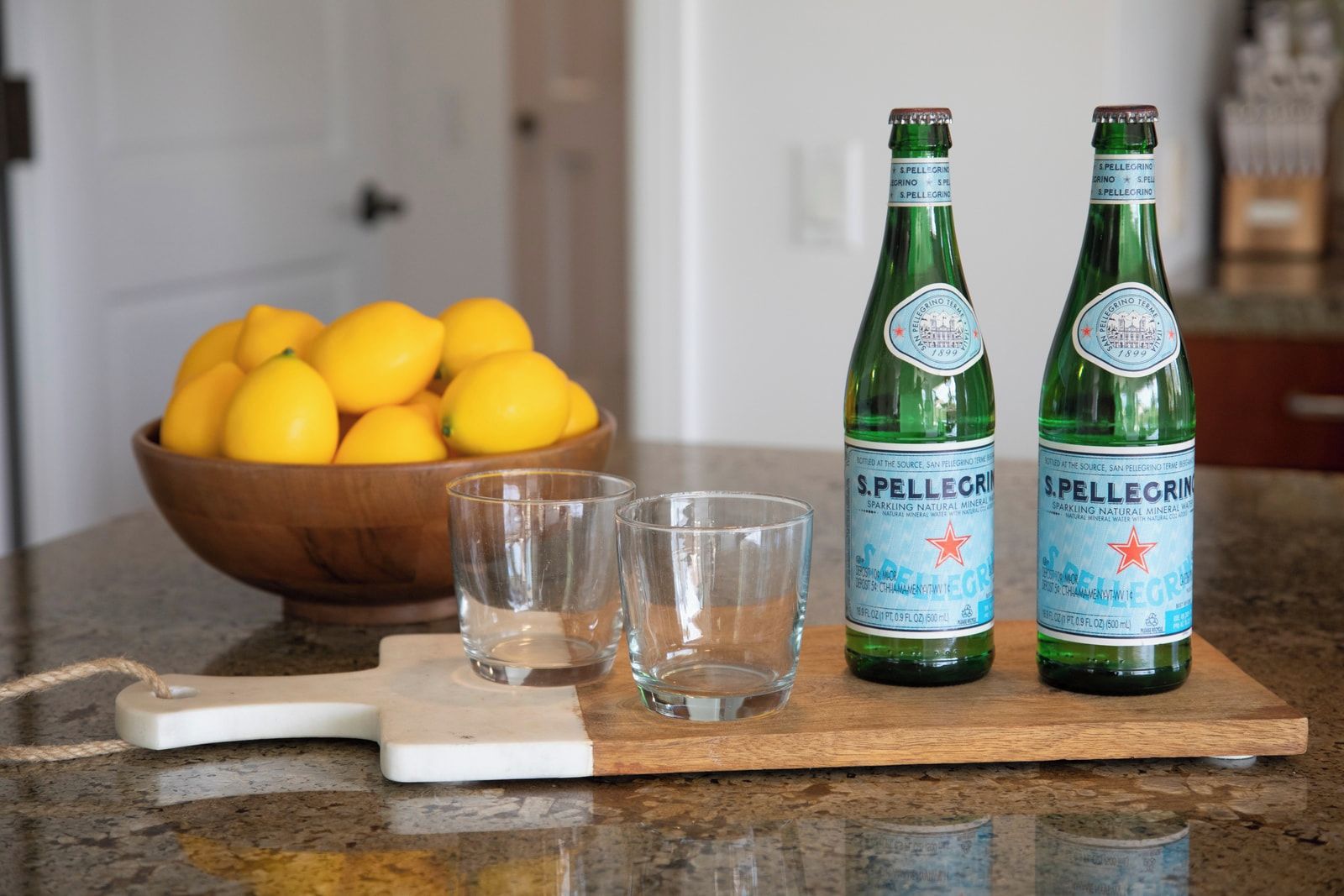 Two bottles of S. Pellegrino sparkling water, a wooden bowl of lemons, and two empty glasses on a kitchen counter.