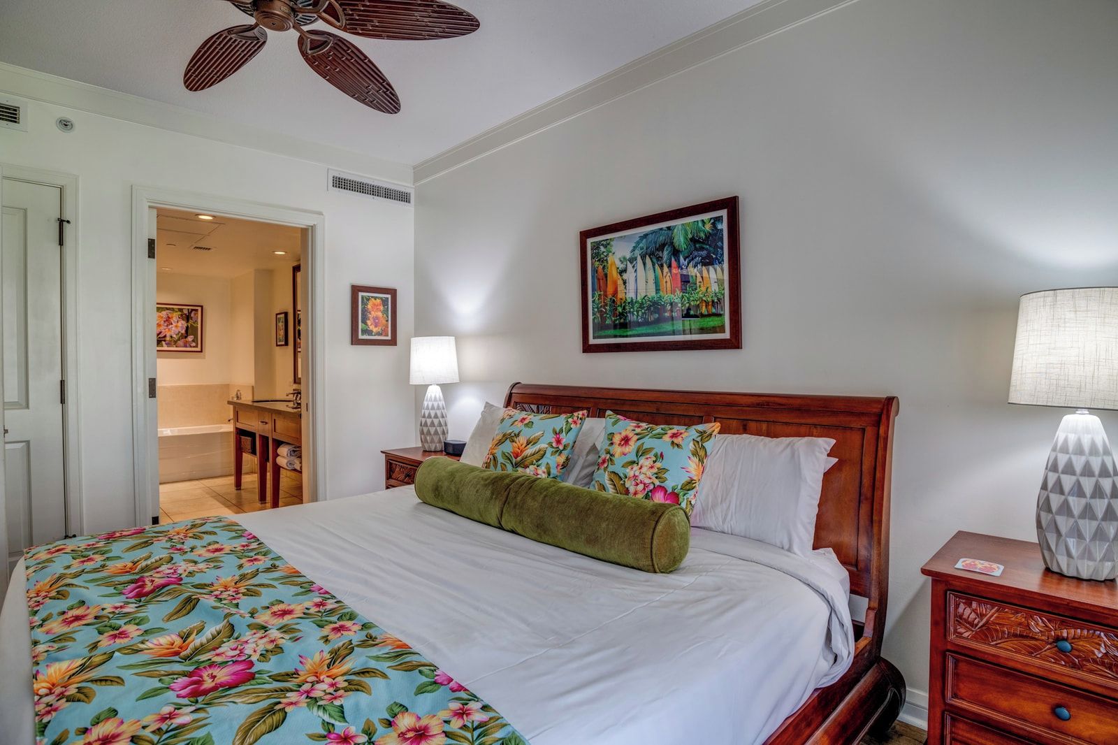 Una habitación de hotel con una cama, cabecera de madera, un cuadro en la pared, una lámpara y una puerta que da al baño.
