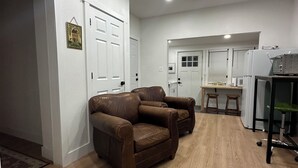 A modern living room with a brown leather sofa, a wooden floor, and a kitchen area with a refrigerator and stools. - Charming 1-bedroom house in perfect Fort Morgan Walking distance to downtown (Fort Morgan)