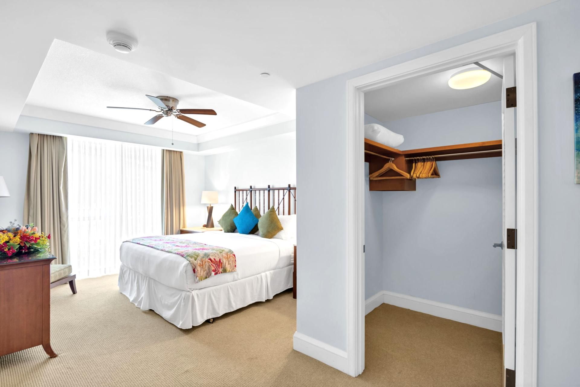 Una habitación de hotel con cama, ventilador de techo, mesita de noche, lámpara, cómoda y armario con ropa colgada.