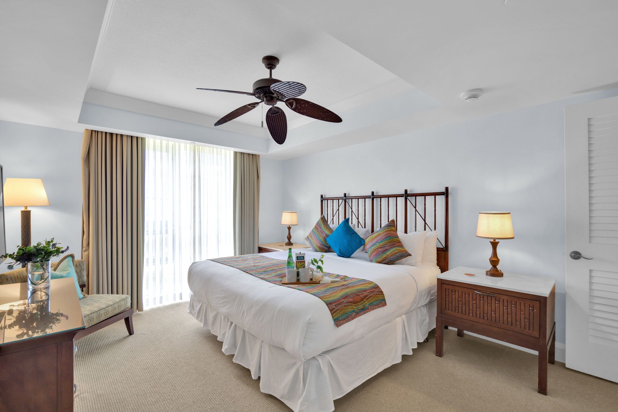 Habitación de hotel con cama, mesitas de noche, escritorio, silla, ventilador de techo y ventana con cortinas.