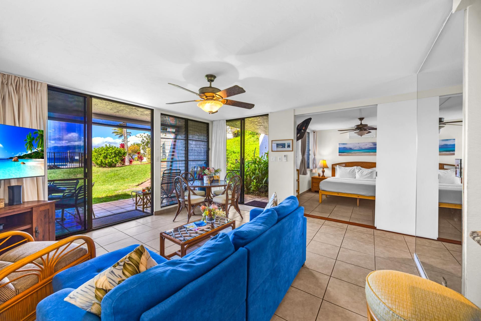 A spacious living area with a blue sofa, a dining table, and chairs. There is a balcony with a view of a tropical landscape.