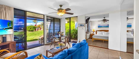 A spacious living area with a blue sofa, a dining table, and chairs. There is a balcony with a view of a tropical landscape.