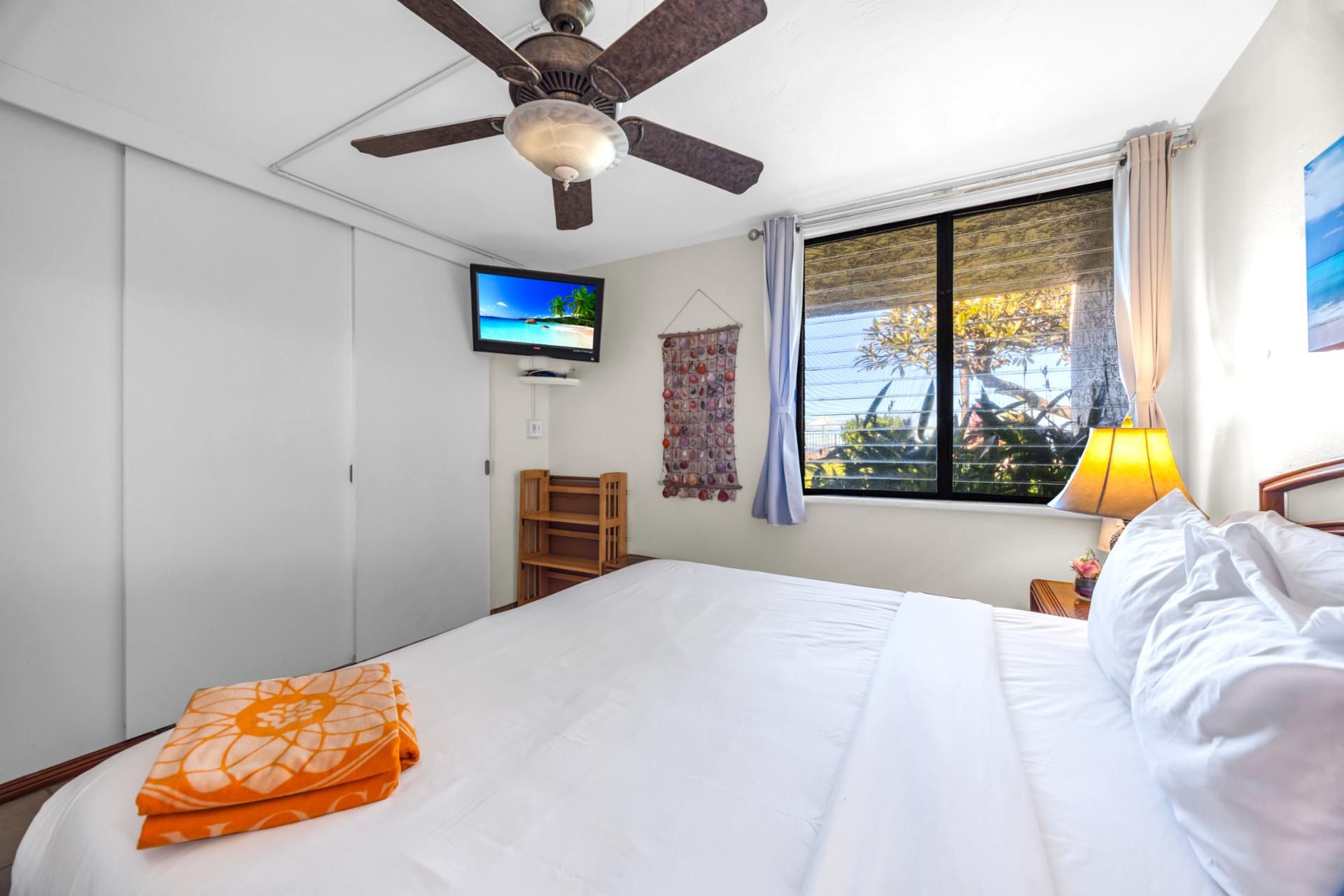Ein Hotelzimmer mit Bett, Deckenventilator, Fernseher, Fenster mit Vorhängen und einem kleinen Tisch mit Lampe.