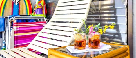 En solig utesittplats med en vit loungestol, en färgglad randig hängmatta och ett glasbord med två drinkar och en blomsteruppsättning.