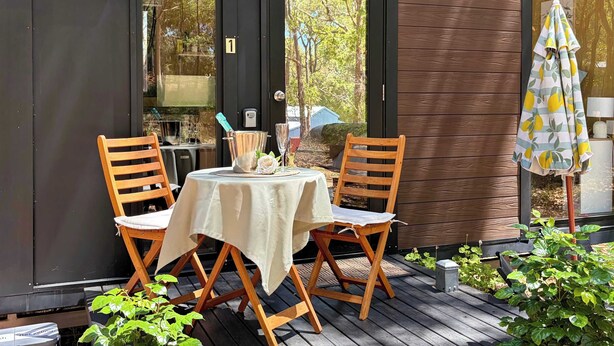 Un espace extérieur aménagé avec deux chaises en bois et une table, une porte vitrée portant un numéro, et un petit parasol.