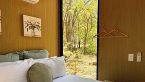 Une chambre à coucher avec un lit, des oreillers, un portant à vêtements et une vue sur les arbres par la fenêtre.
