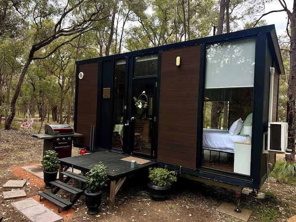A compact, modern cabin with a glass door leading to a bedroom, surrounded by a natural forest setting.